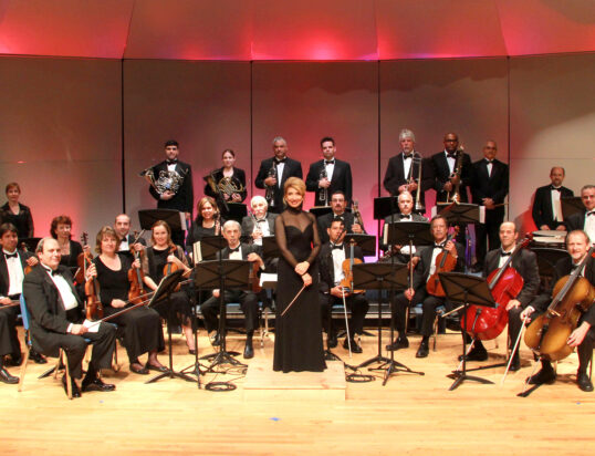 Thumbnail of http://An%20orchestra%20on%20stage,%20members%20seated%20with%20instruments%20like%20violins%20and%20cellos,%20conductor%20in%20black%20dress%20stands%20in%20front.%20Warm,%20elegant%20ambiance.