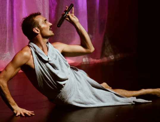 Thumbnail of http://A%20man%20wrapped%20in%20a%20grey%20towel%20reclines%20dramatically%20on%20stage,%20pretending%20to%20sing%20into%20a%20hairbrush.%20The%20backdrop%20is%20illuminated%20in%20pink,%20creating%20an%20artistic,%20theatrical%20atmosphere.