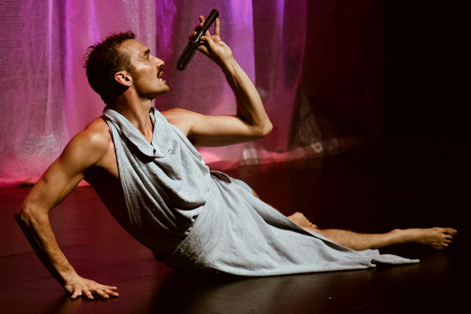 A man wrapped in a grey towel reclines dramatically on stage, pretending to sing into a hairbrush. The backdrop is illuminated in pink, creating an artistic, theatrical atmosphere.