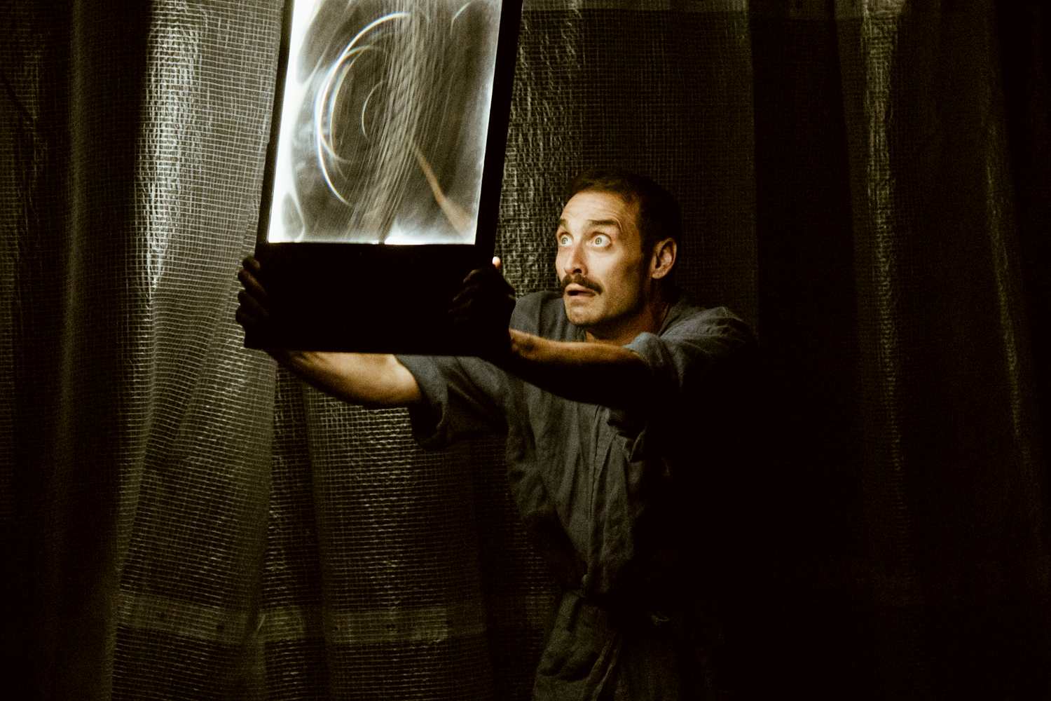 A man gazes upwards, holding a glowing, swirling light box. He stands against a dark, textured curtain, creating a dramatic and mysterious atmosphere.
