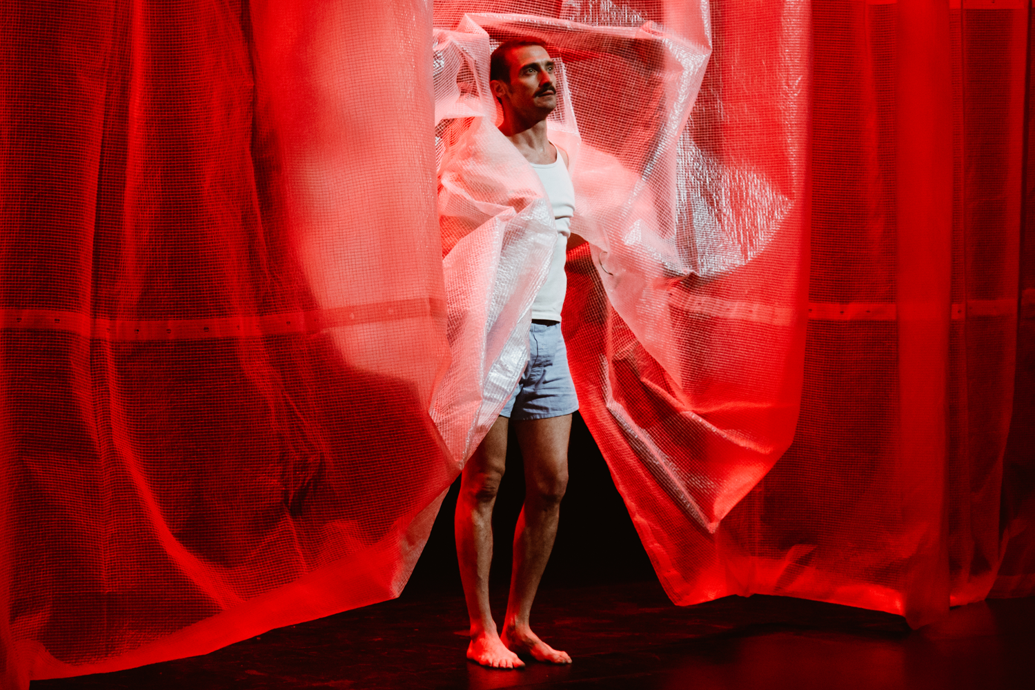 A man in shorts and a tank top stands barefoot, emerging through red transparent curtains. The scene is dramatically lit, casting a bold, theatrical mood.