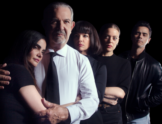 Thumbnail of http://A%20group%20of%20five%20people%20stands%20together%20against%20a%20black%20background.%20The%20central%20figure%20is%20an%20older%20man%20in%20a%20white%20shirt%20and%20black%20tie,%20with%20a%20serious%20expression,%20gently%20embracing%20a%20woman%20to%20his%20right.%20The%20others,%20dressed%20in%20black,%20stand%20closely%20in%20a%20line,%20exuding%20a%20tense,%20dramatic%20mood.