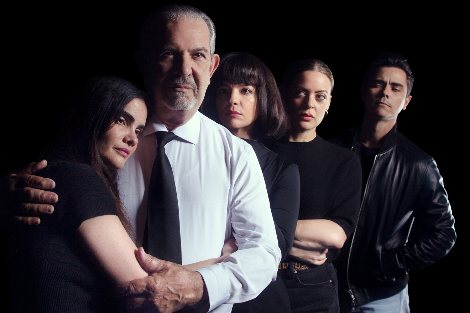A group of five people stands together against a black background. The central figure is an older man in a white shirt and black tie, with a serious expression, gently embracing a woman to his right. The others, dressed in black, stand closely in a line, exuding a tense, dramatic mood.
