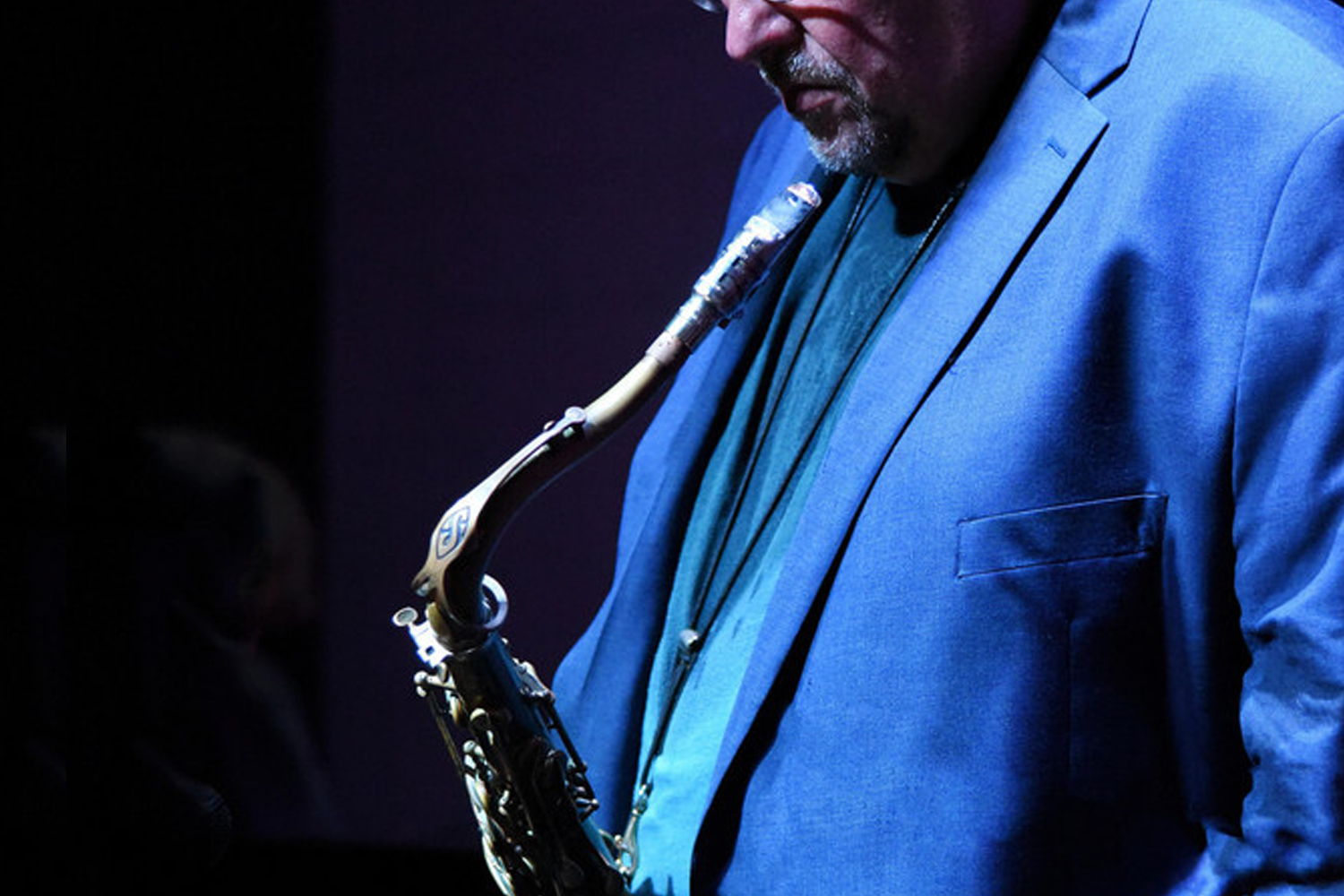 Man in a blue blazer playing a saxophone under soft lighting, focused and introspective. The atmosphere feels calm and intimate.