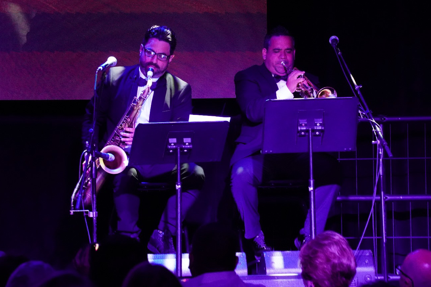 Two men in suits performing on stage, one playing a saxophone and the other playing a trumpet, creating a lively musical atmosphere before an audience.