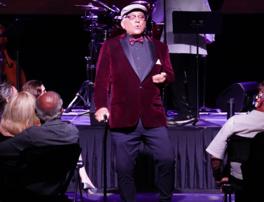 Thumbnail of http://A%20man%20in%20a%20red%20velvet%20suit,%20bow%20tie%20and%20hat%20on%20stage%20with%20the%20audience%20watching%20him%20in%20the%20foreground.