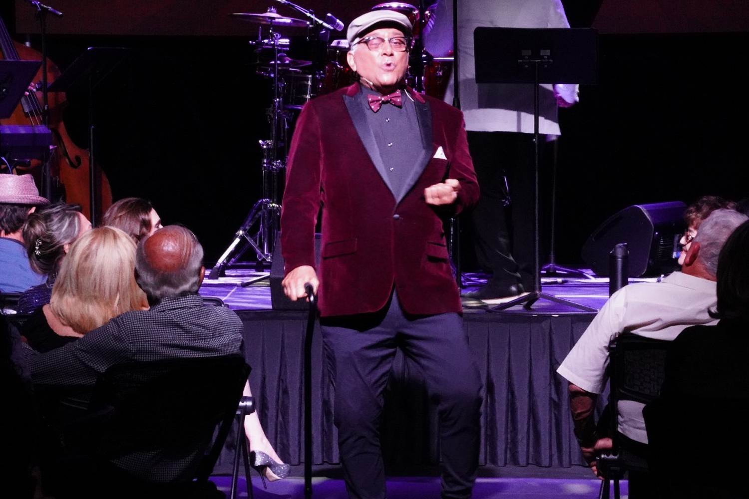 A man in a red velvet suit, bow tie and hat on stage with the audience watching him in the foreground.