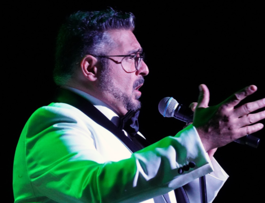 Thumbnail of http://A%20man%20in%20glasses%20passionately%20sings%20into%20a%20microphone%20on%20stage,%20wearing%20a%20white%20suit%20with%20a%20black%20bow%20tie,%20illuminated%20by%20dramatic%20lighting.