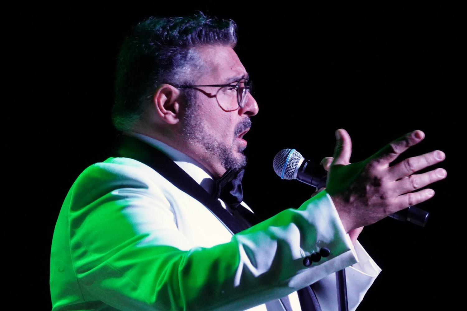 A man in glasses passionately sings into a microphone on stage, wearing a white suit with a black bow tie, illuminated by dramatic lighting.