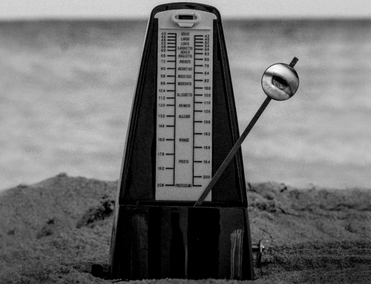 Thumbnail of http://Black-and-white%20image%20of%20a%20metronome%20on%20sand%20near%20the%20ocean.%20The%20metronome's%20pendulum%20has%20an%20eye%20illustration.