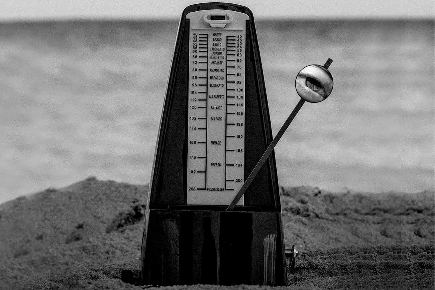 Black-and-white image of a metronome on sand near the ocean. The metronome's pendulum has an eye illustration.
